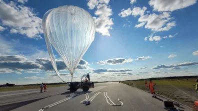 Un homme conduit un véhicule retenant un ballon au sol. Au loin, plusieurs personnes autour du véhicule.